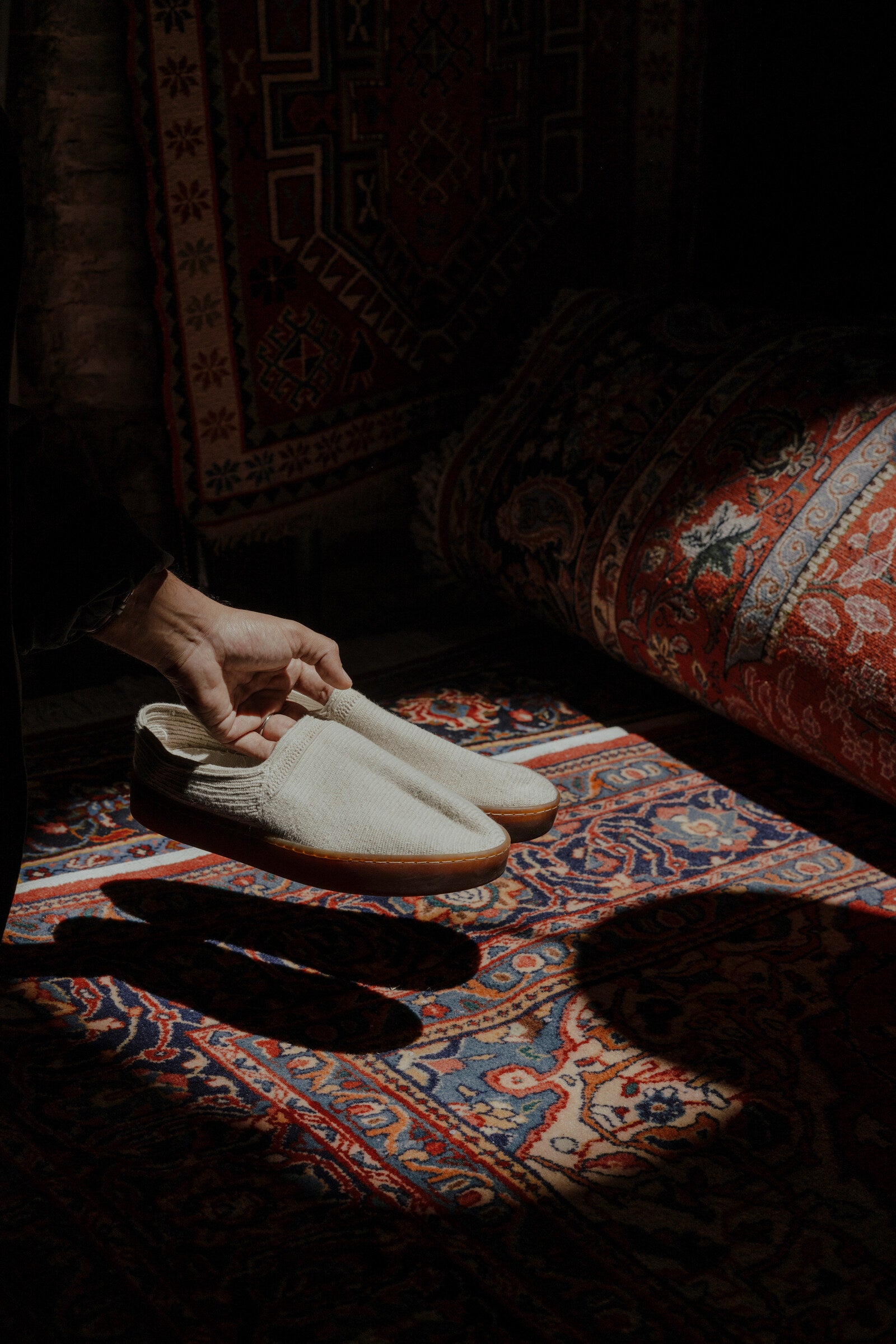 Une main dépose une paire de Pâpouches sur des tapis persans, dans un clair-obscur
